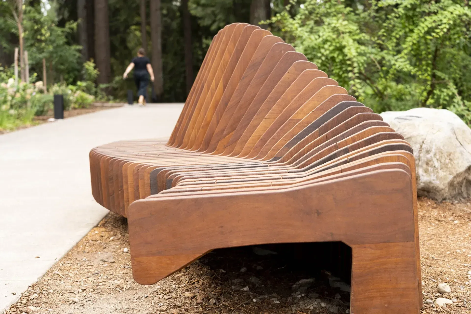 Wooden bench in a park setting with a person walking on a path in the background. 
