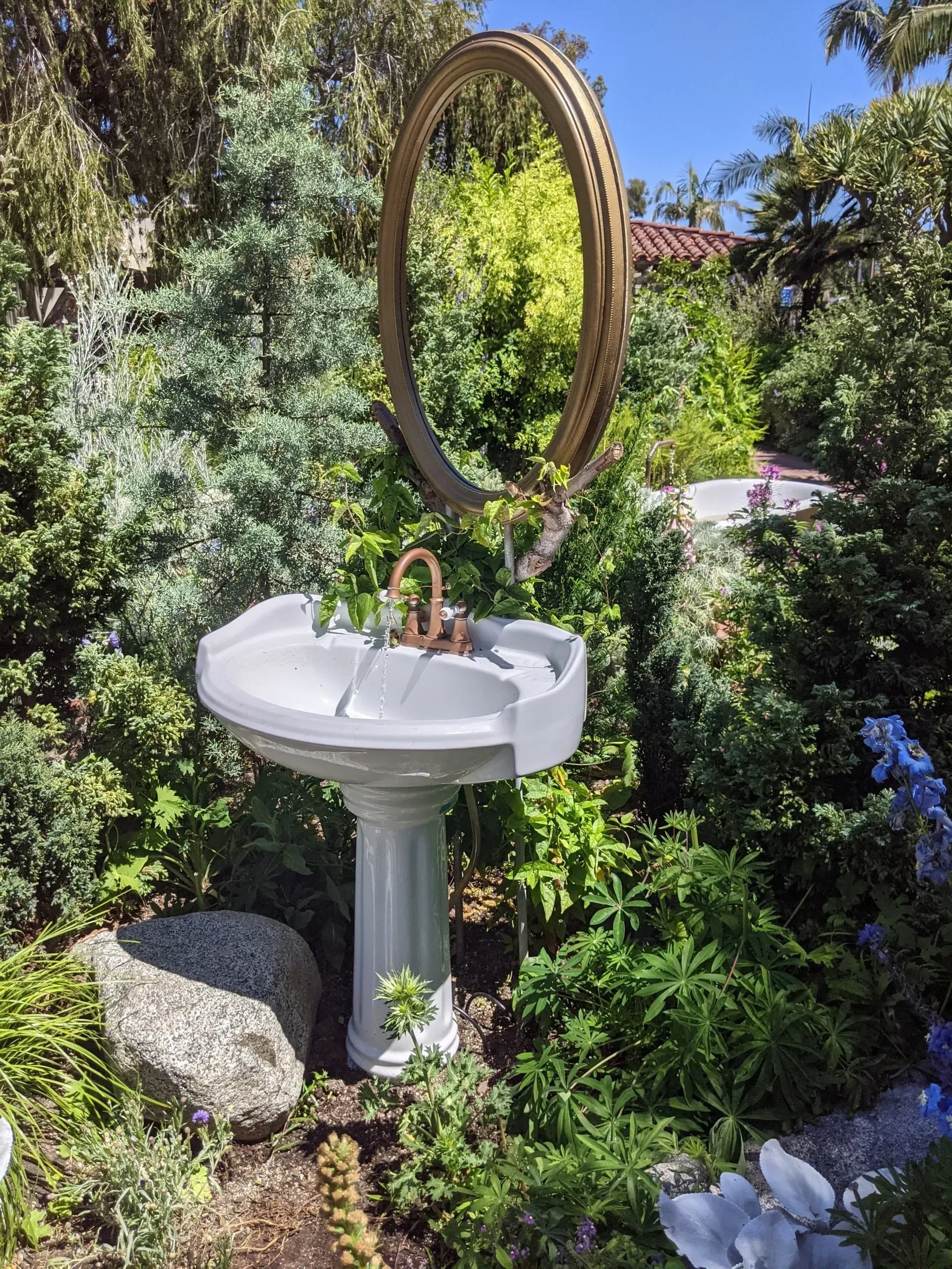 A pedestal sink with a cracked basin in a lush garden, adorned with a golden, oval mirror.
