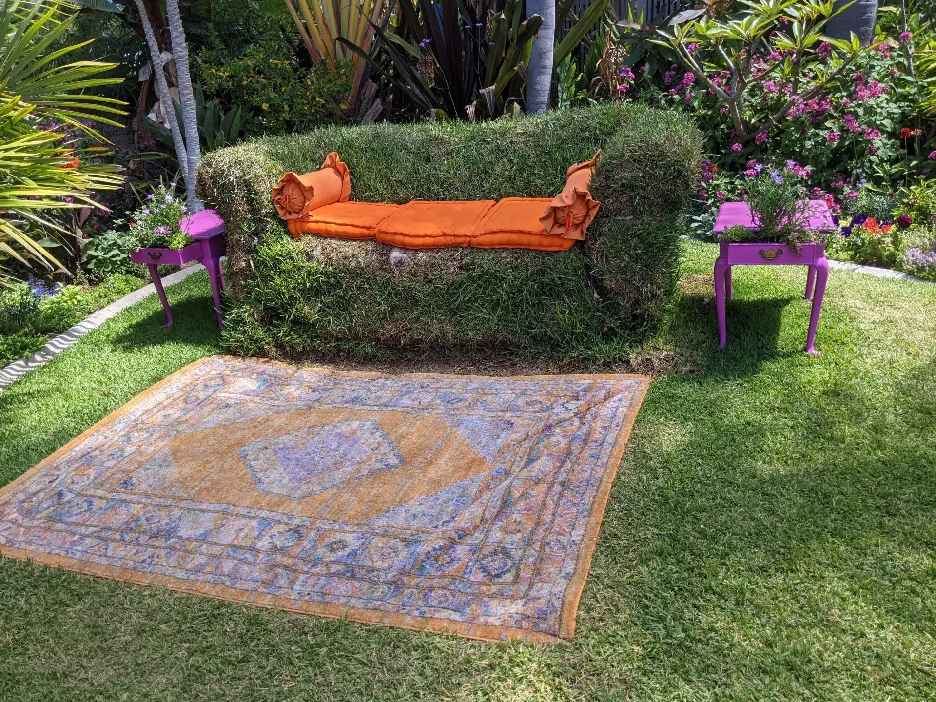 Hedge chair with orange cushions, flanked by purple tables, and a patterned rug on a grassy lawn.