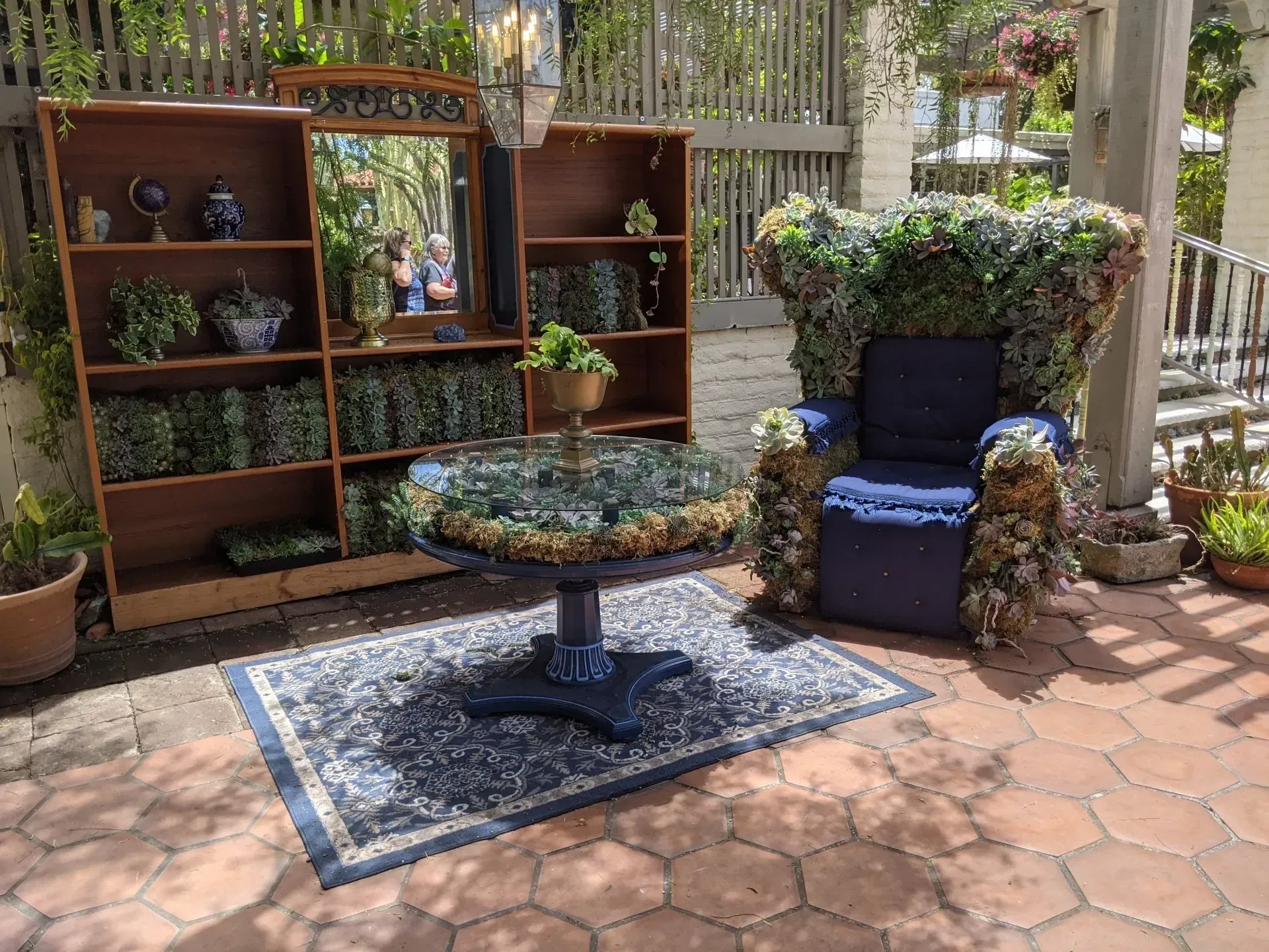 Outdoor garden scene with a floral-covered armchair, table, and bookshelf. 