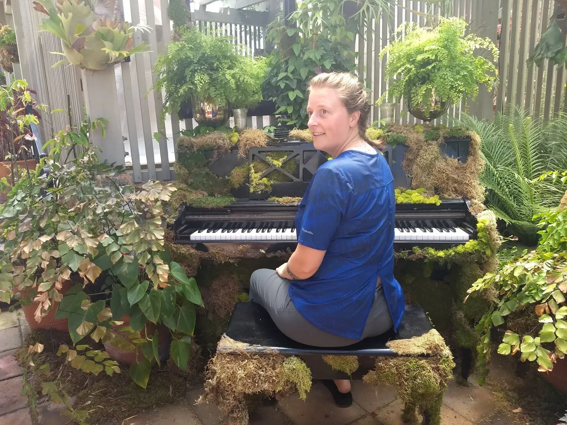 Woman sitting at a piano covered in moss and plants in a garden setting. She smiles, wearing a blue shirt and gray pants.