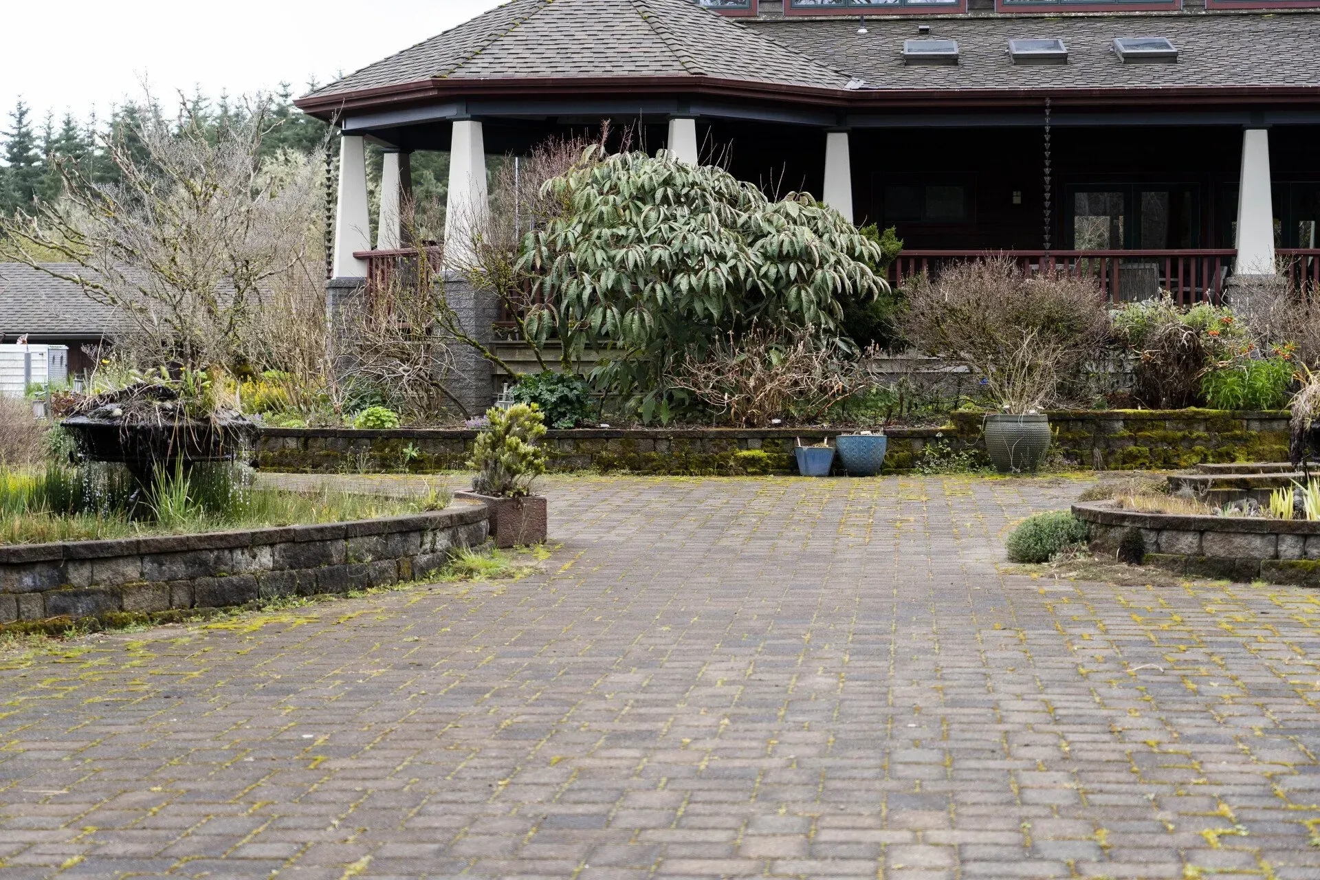 Brick driveway leading to a house with a wraparound porch and garden beds. The house has columns and a dark roof.