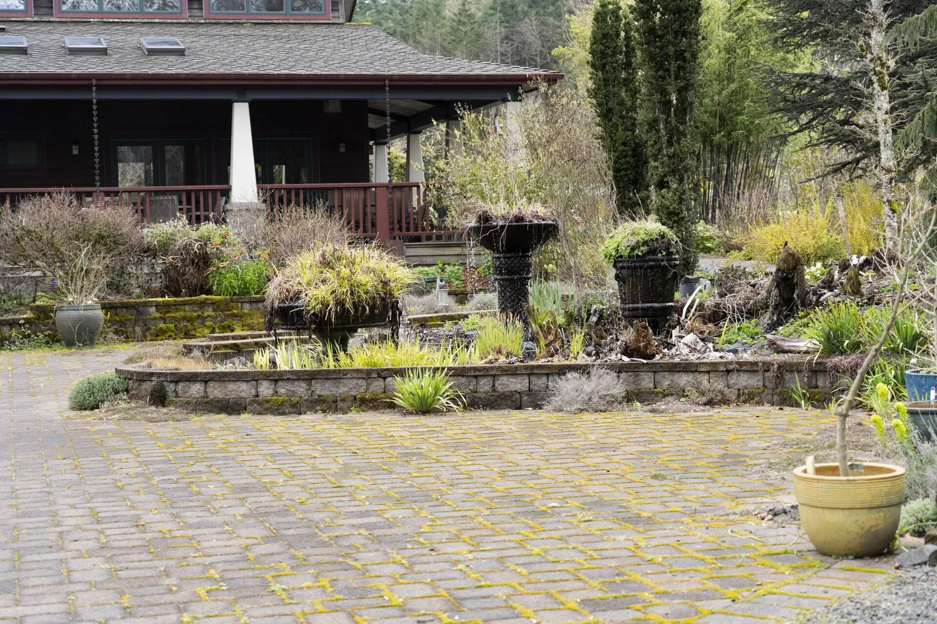 A brick-paved driveway leads to a house with a porch and a stone planter with greenery. A potted plant is in the foreground.