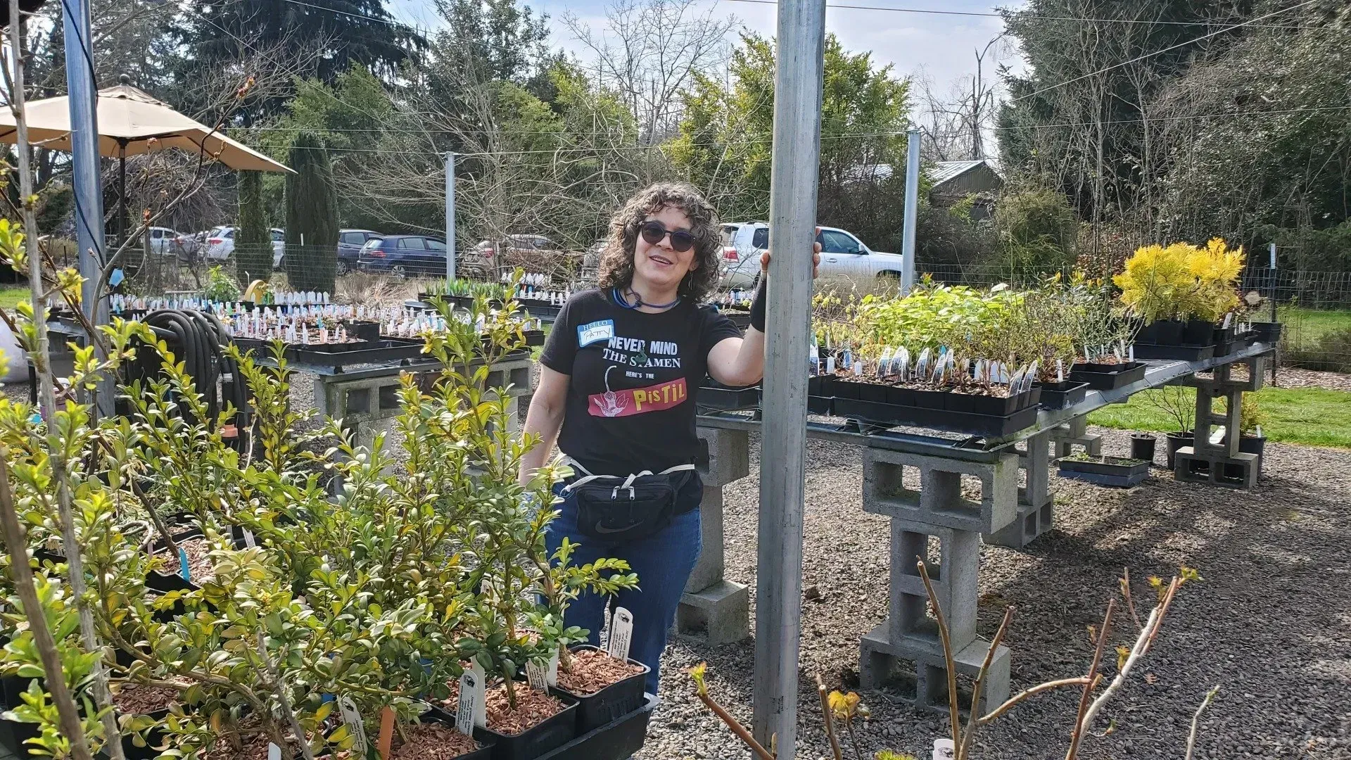 Woman smiling, standing in a nursery surrounded by potted plants. She wears sunglasses and a t-shirt.