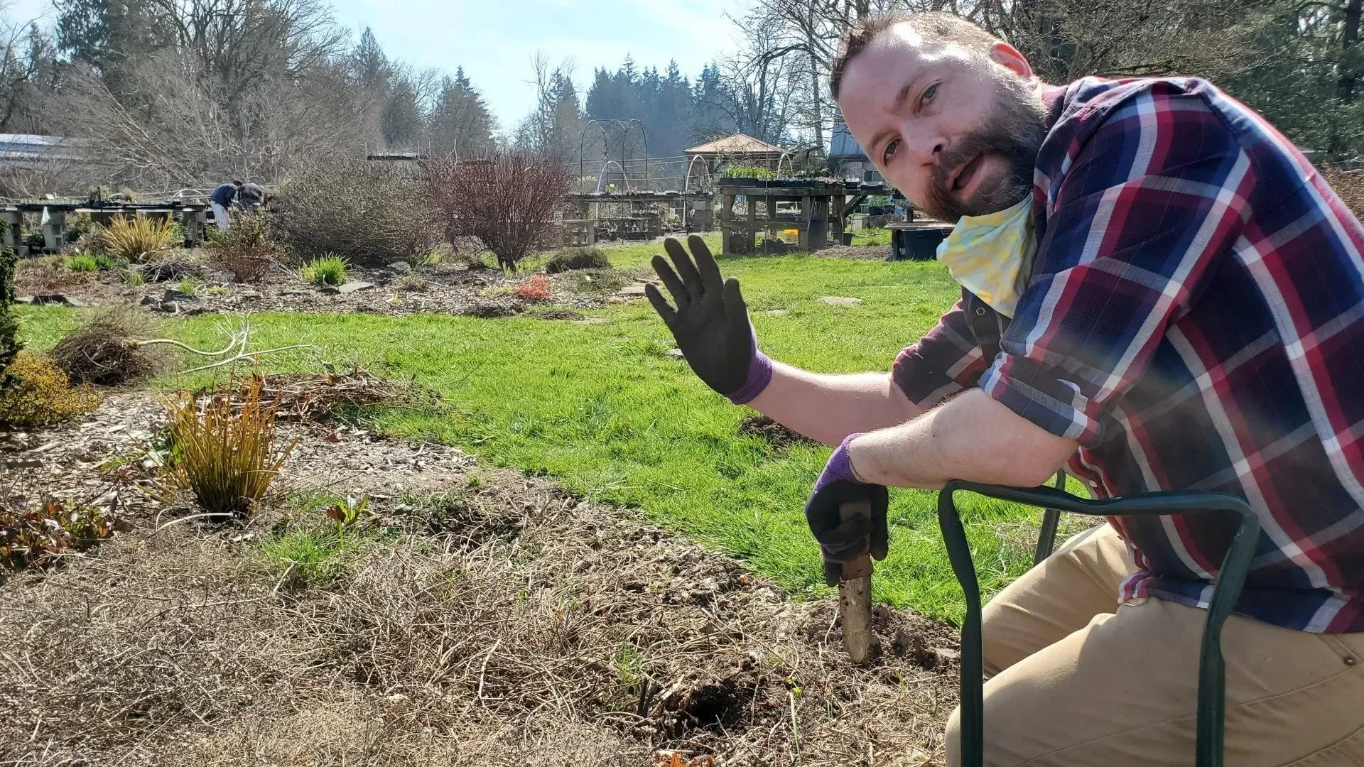 Man in a garden waves while wearing gloves and a mask. The ground in front is brown, and grass grows beyond.