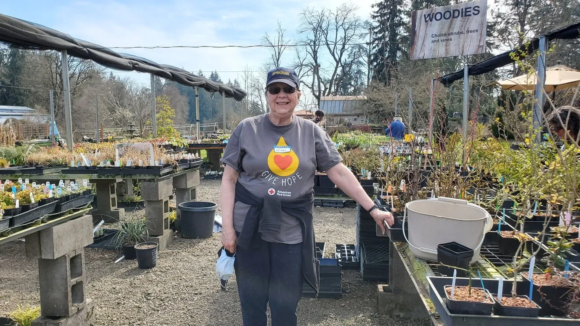 A woman in a gray shirt stands in a plant nursery. Tables of potted plants surround her; the setting appears outdoors.
