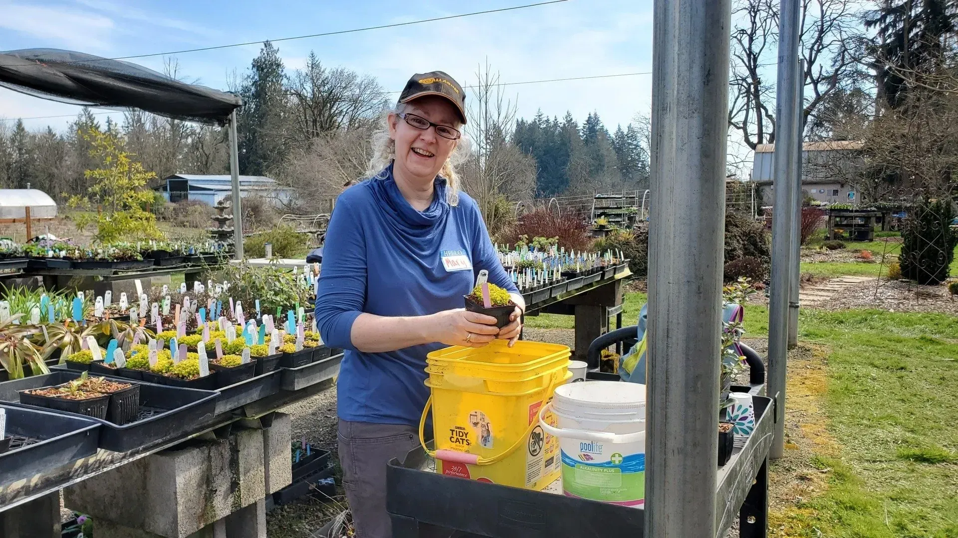 A woman in a garden center wearing a hat and glasses smiles while working with plants near buckets.