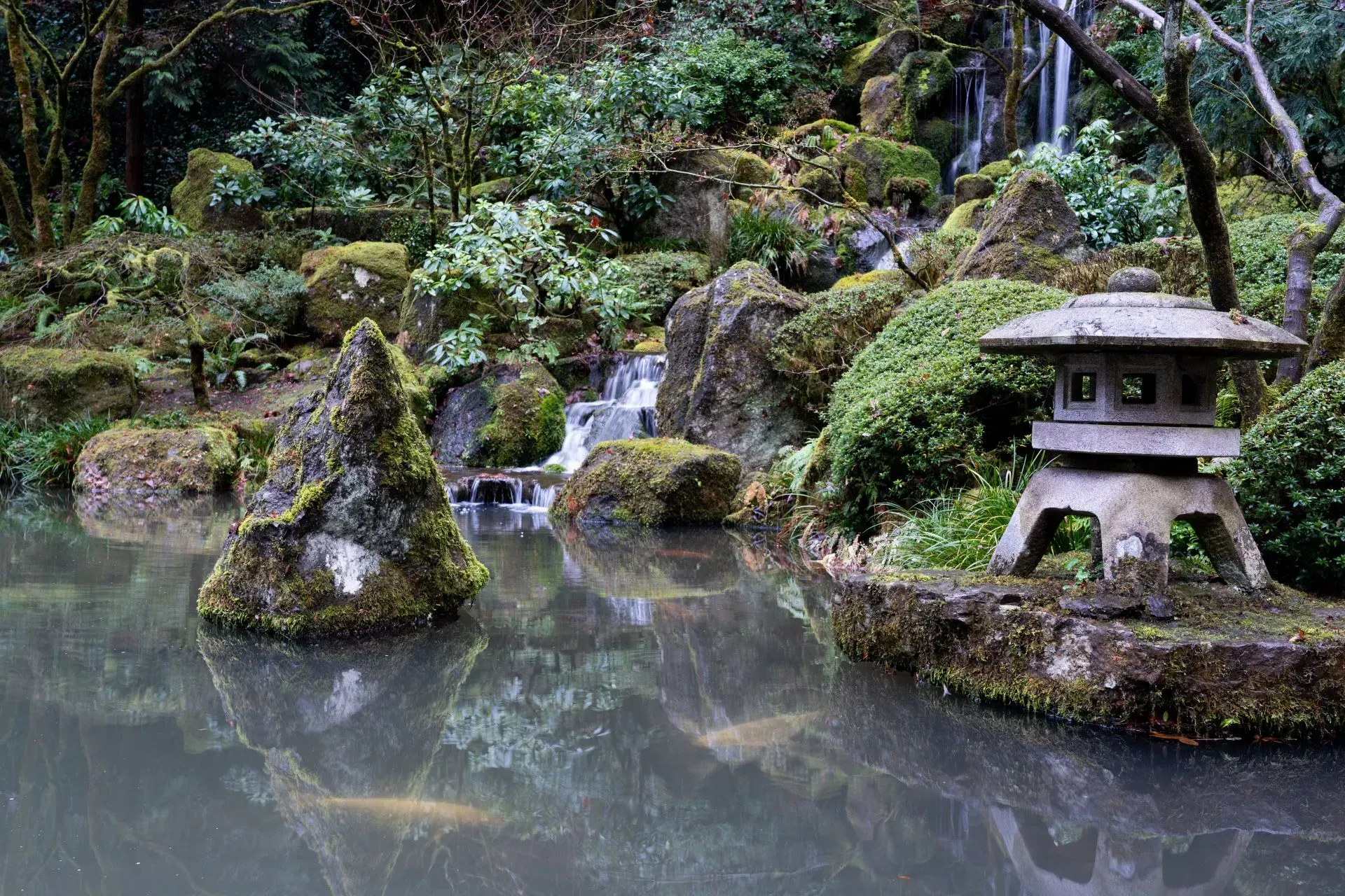 A Japanese garden with a pond, rocky landscape, stone lantern, and small waterfall.