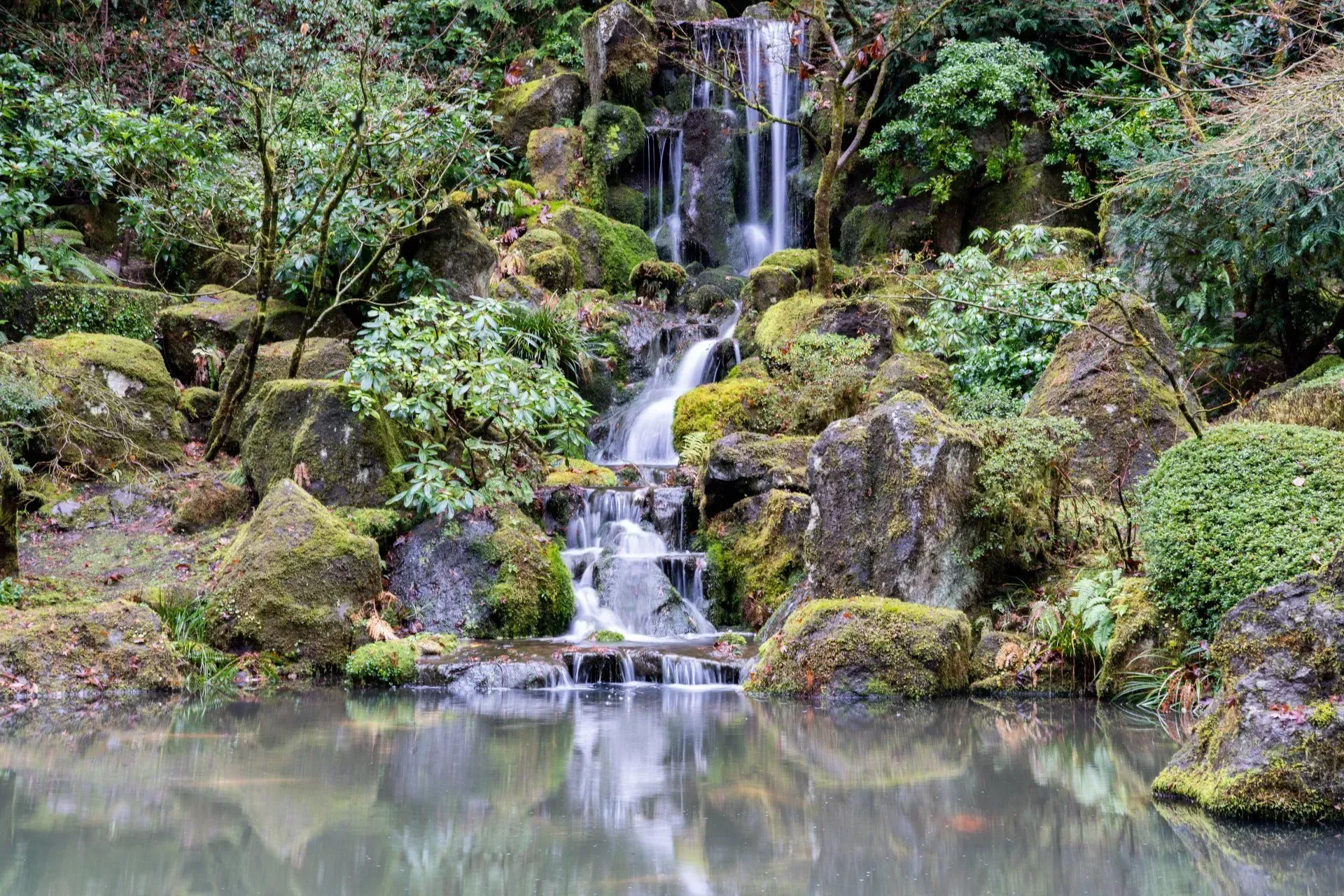 A small waterfall cascades into a pond surrounded by moss-covered rocks and green foliage in a garden setting.