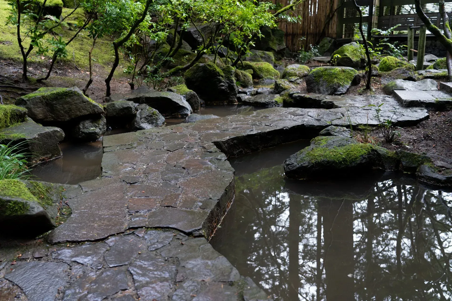 A moss-covered stone bridge over a pond in a Japanese garden. Reflections of trees are visible in the still water.