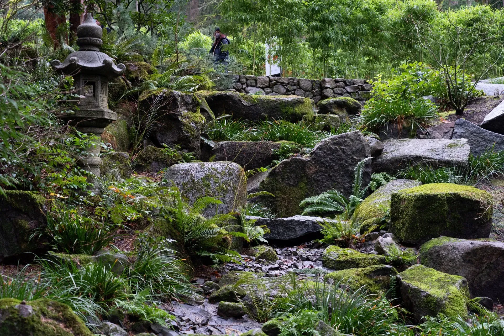 Stone steps lead through a moss-covered garden with a stone lantern and people visible in the distance. 