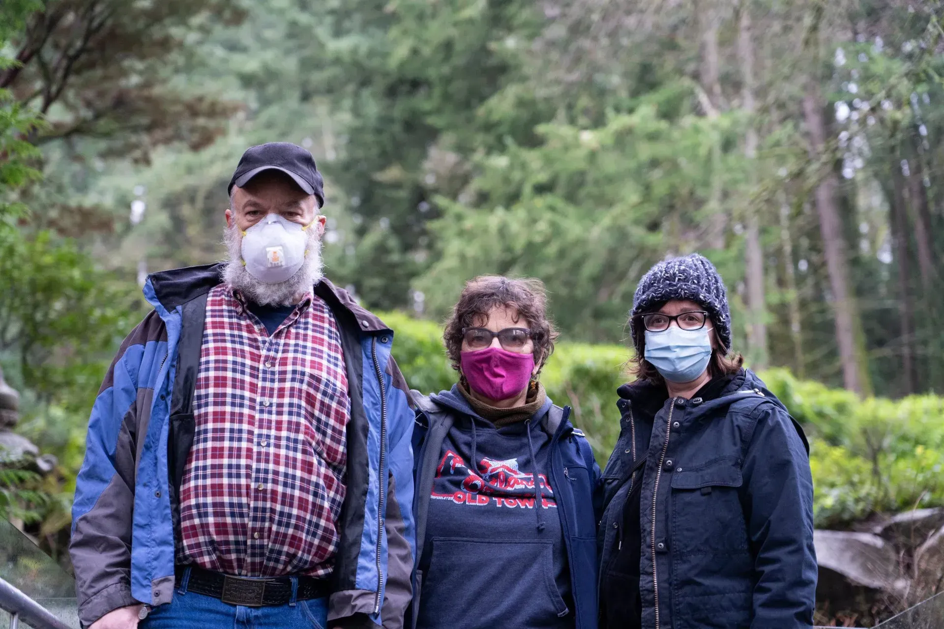 Three people in jackets and face masks stand outdoors in a wooded area. The man on the left has a long white beard.