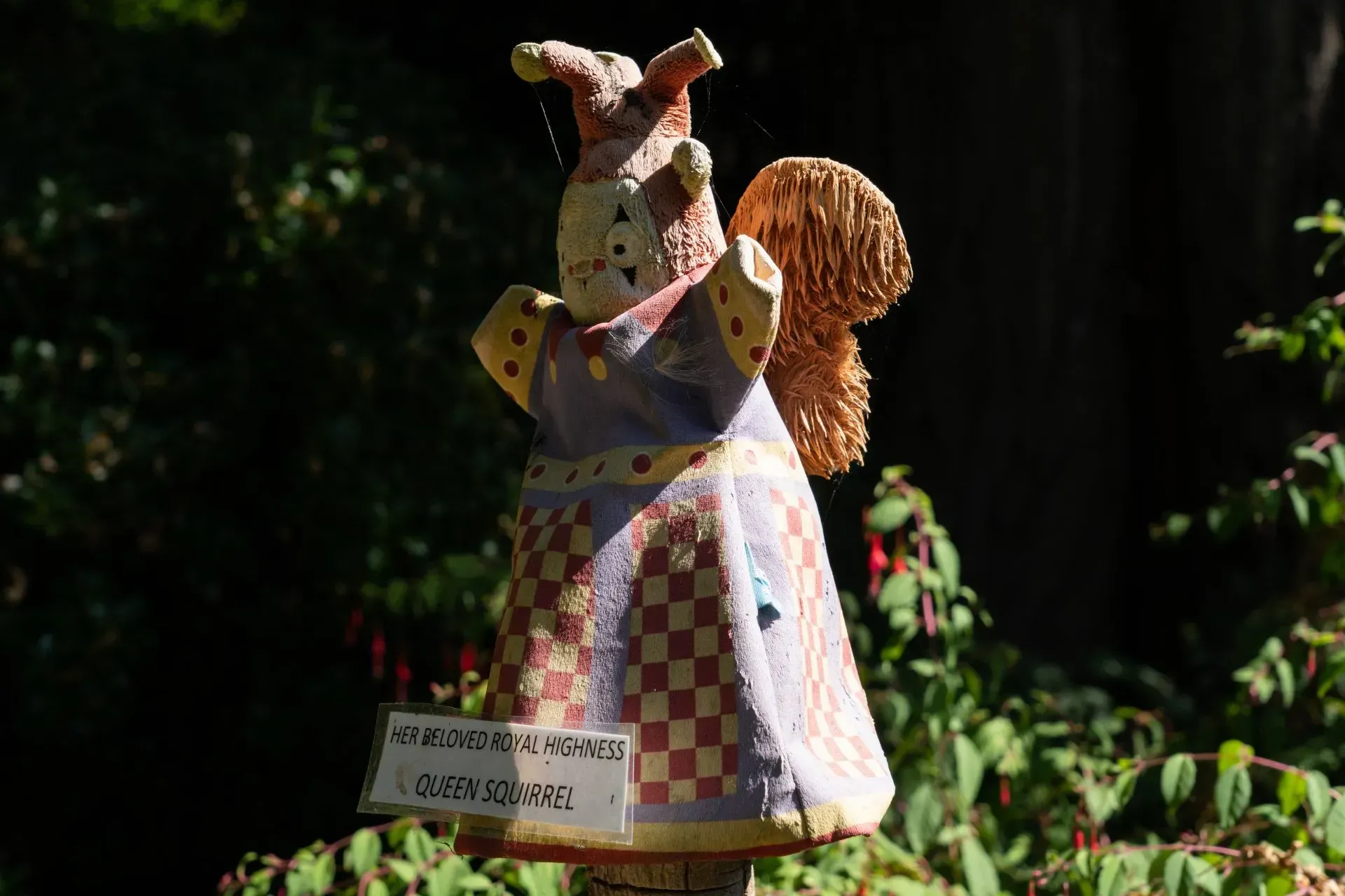 A whimsical, ceramic angel-like figure in a purple and brown dress stands on a wooden post.