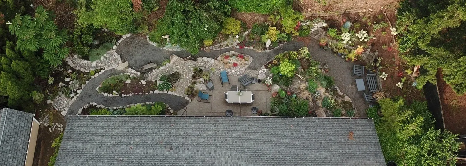 Overhead view of a backyard garden with a stone path winding through greenery and leading to a patio with a table and chairs.