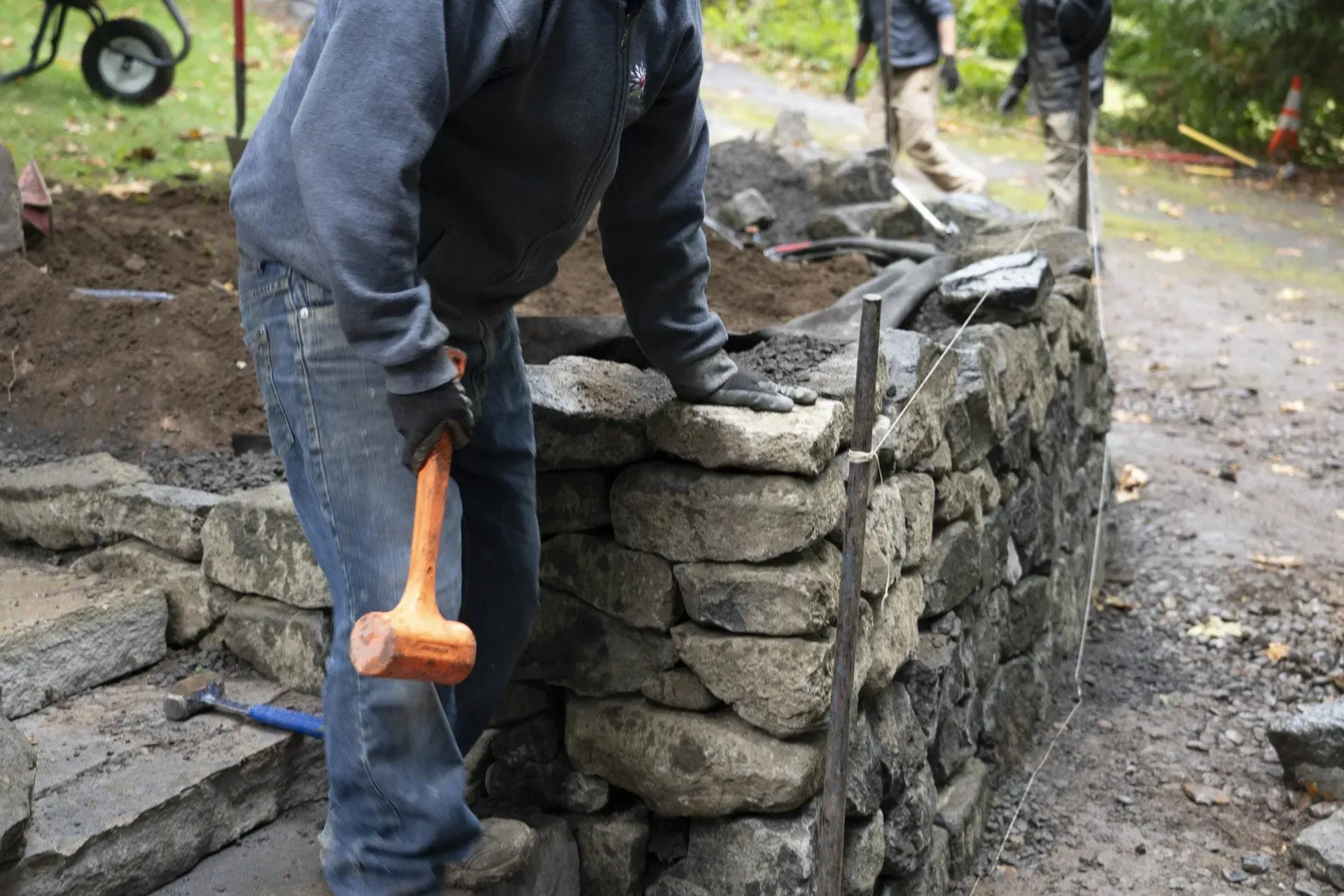 A person in blue jeans and gloves uses a rubber mallet to work on a stone wall outdoors.