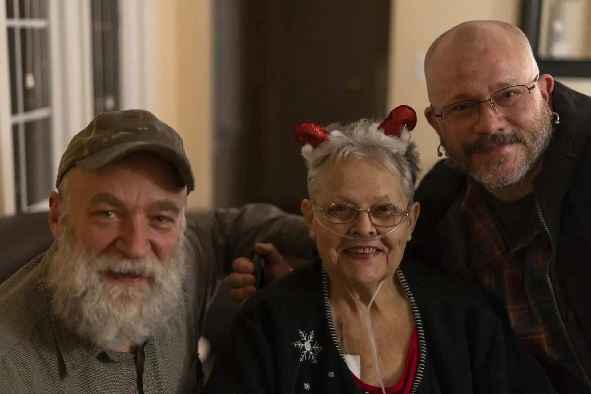 Three people smiling: an elderly woman wearing a festive headband sits between two men with beards. Indoors, warm lighting.