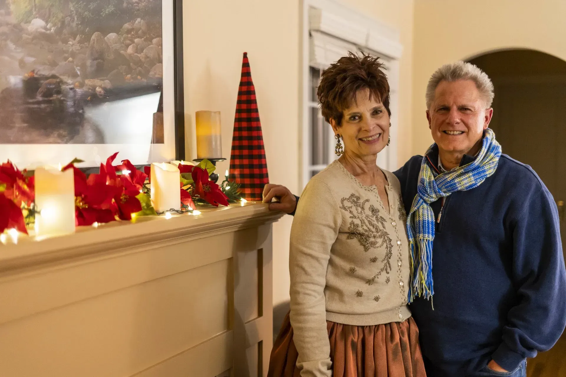 A smiling couple stands next to a fireplace decorated for the holidays.
