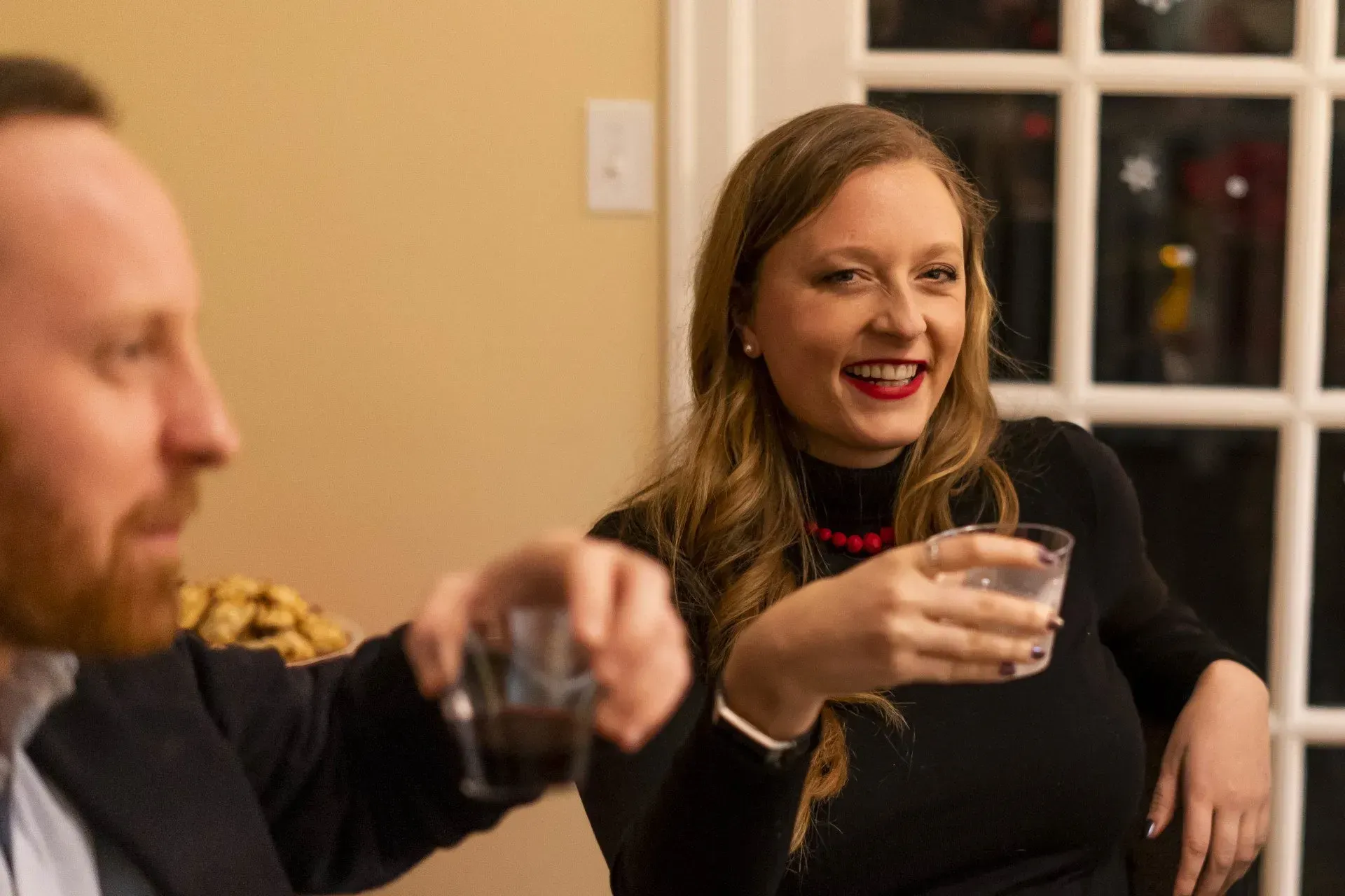Woman with red lipstick smiles, holding a glass. A man beside her raises his glass. They are indoors.