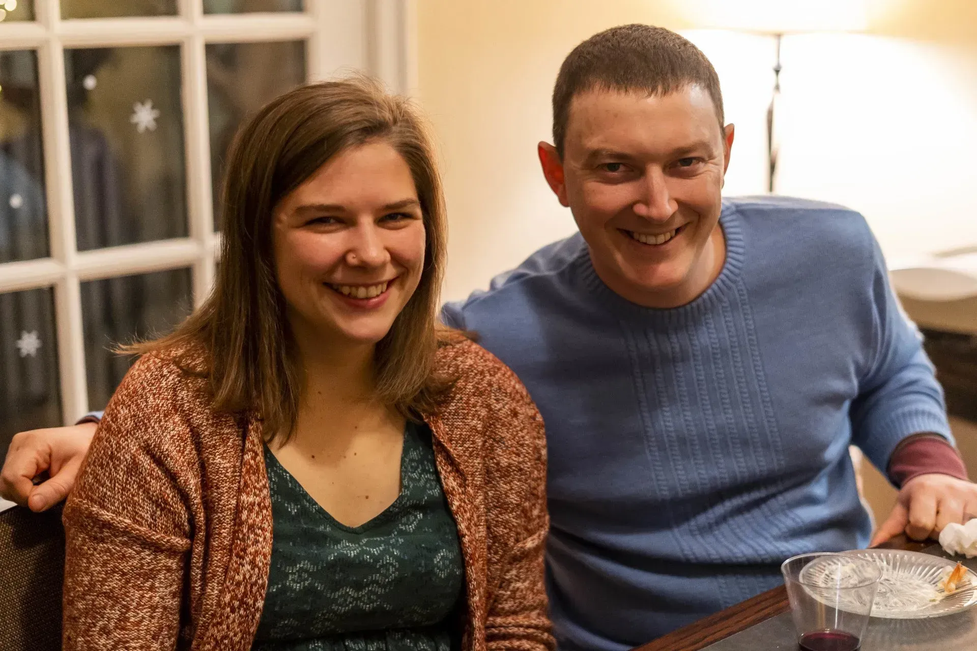 A smiling man and woman sit at a table. The woman wears a brown cardigan and green top; the man wears a blue sweater.