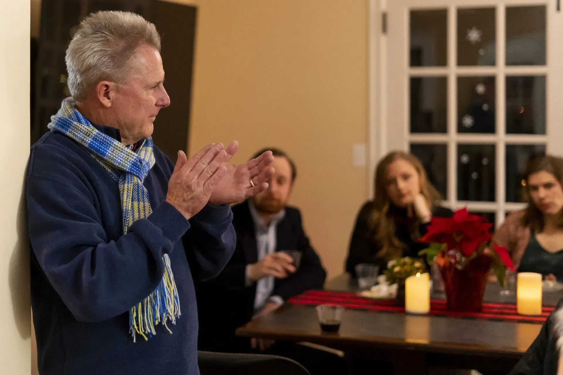 Man with silver hair and blue scarf speaks to a group at a table. The setting appears to be a living room or dining room.