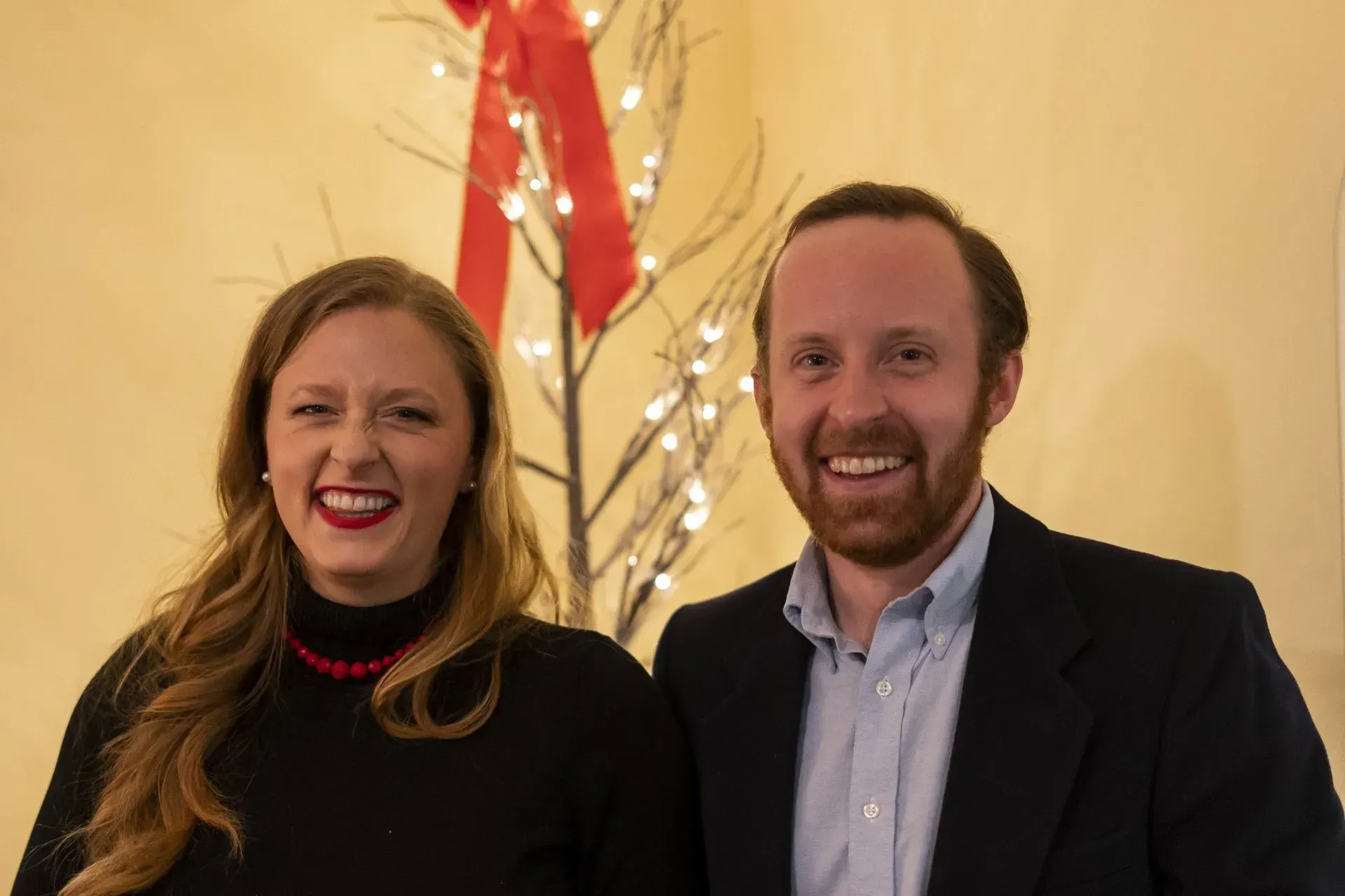 Woman and man smiling, posing together in front of a decorated tree with red bow.