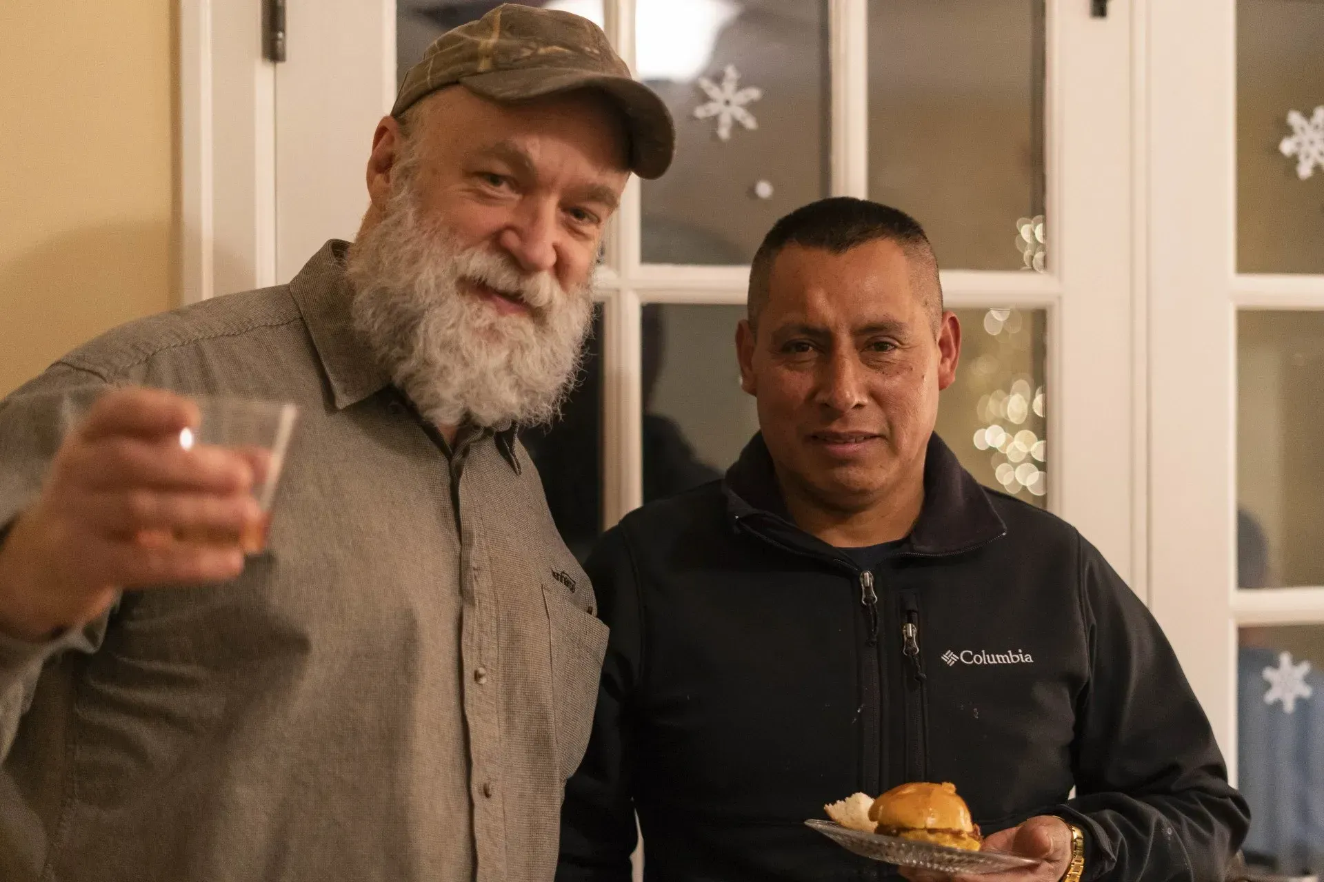 Two men smiling at a party; one holding a drink, the other a plate of food.