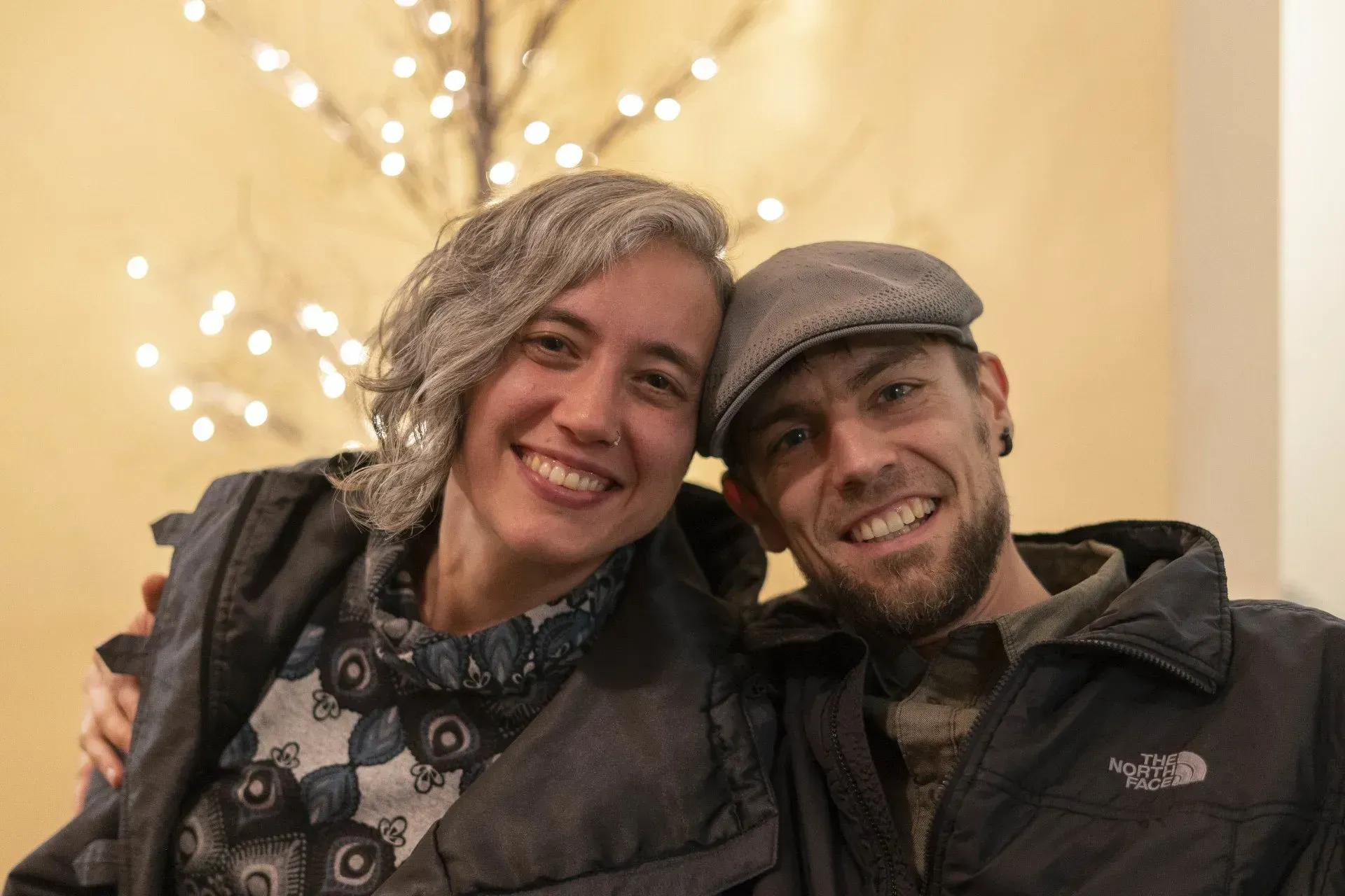 Woman with gray curly hair and man in a newsboy cap smile, embracing. Background includes a lit-up branch.
