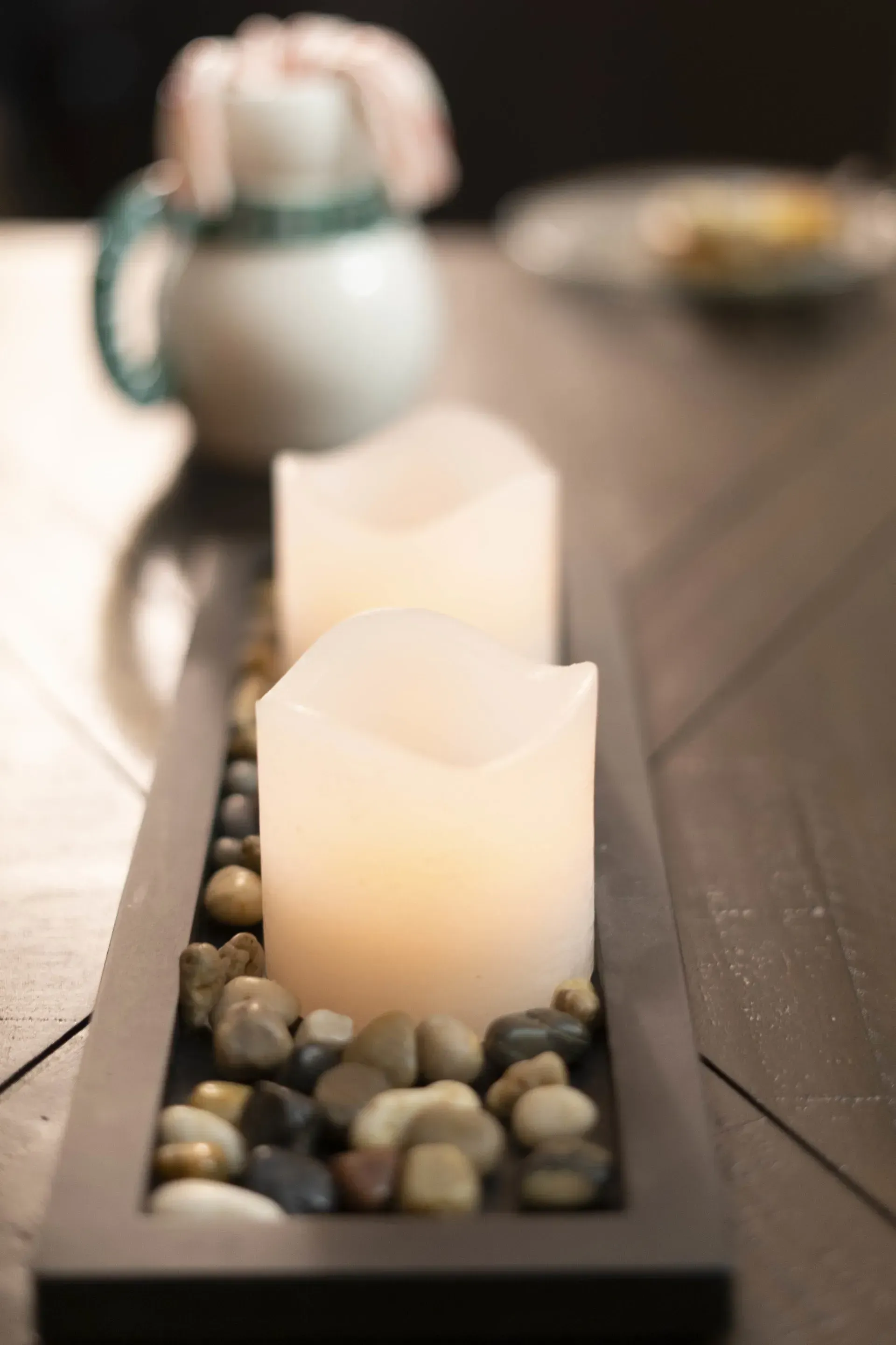 Two lit, white candles in a wooden tray filled with pebbles on a dark wooden table, a teapot is in the background.