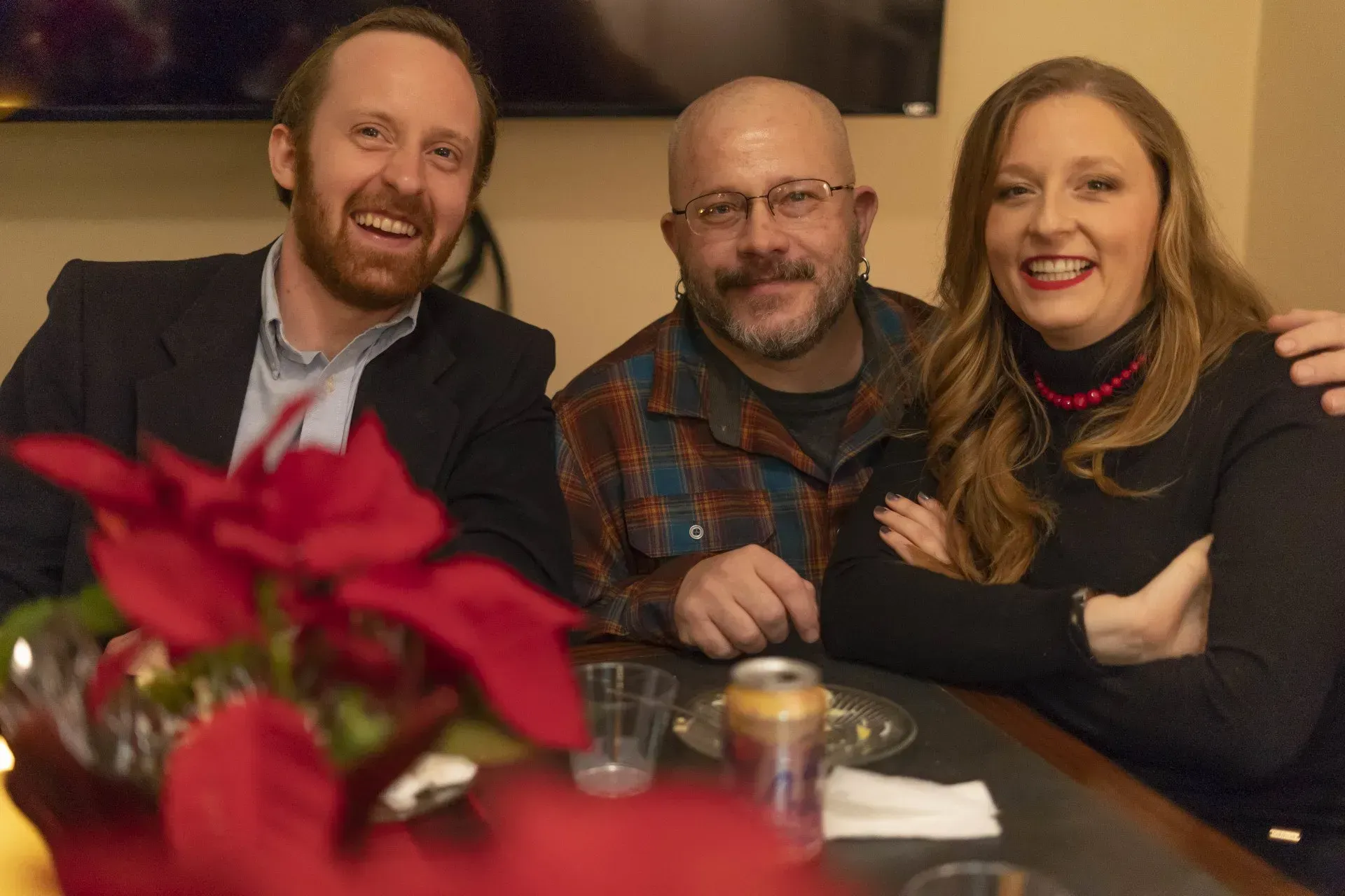 Three smiling people gathered around a table with red poinsettia flowers, a beer can, and glassware.