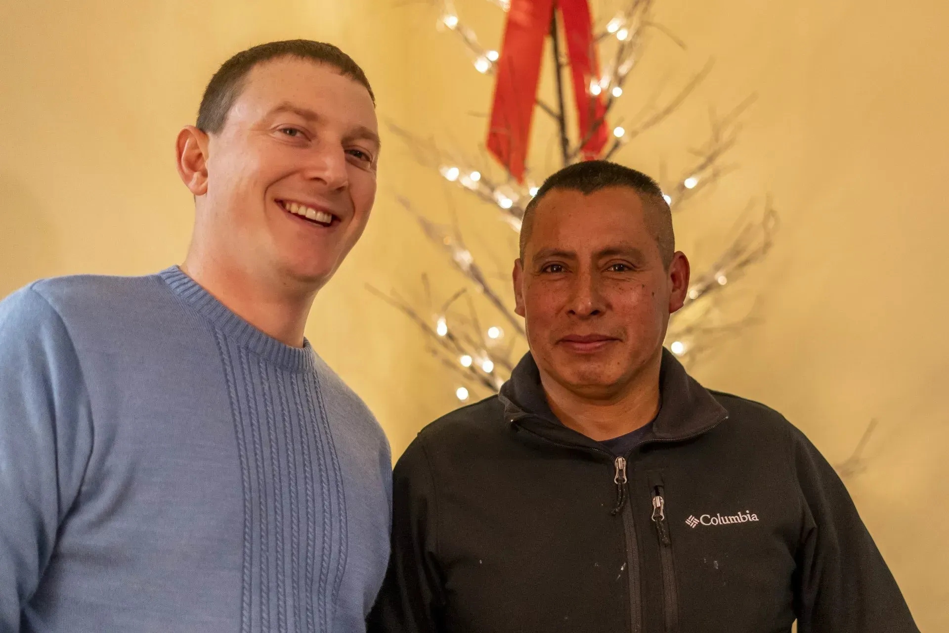 Two men smiling, standing in front of a decorative light display with a red bow. 
