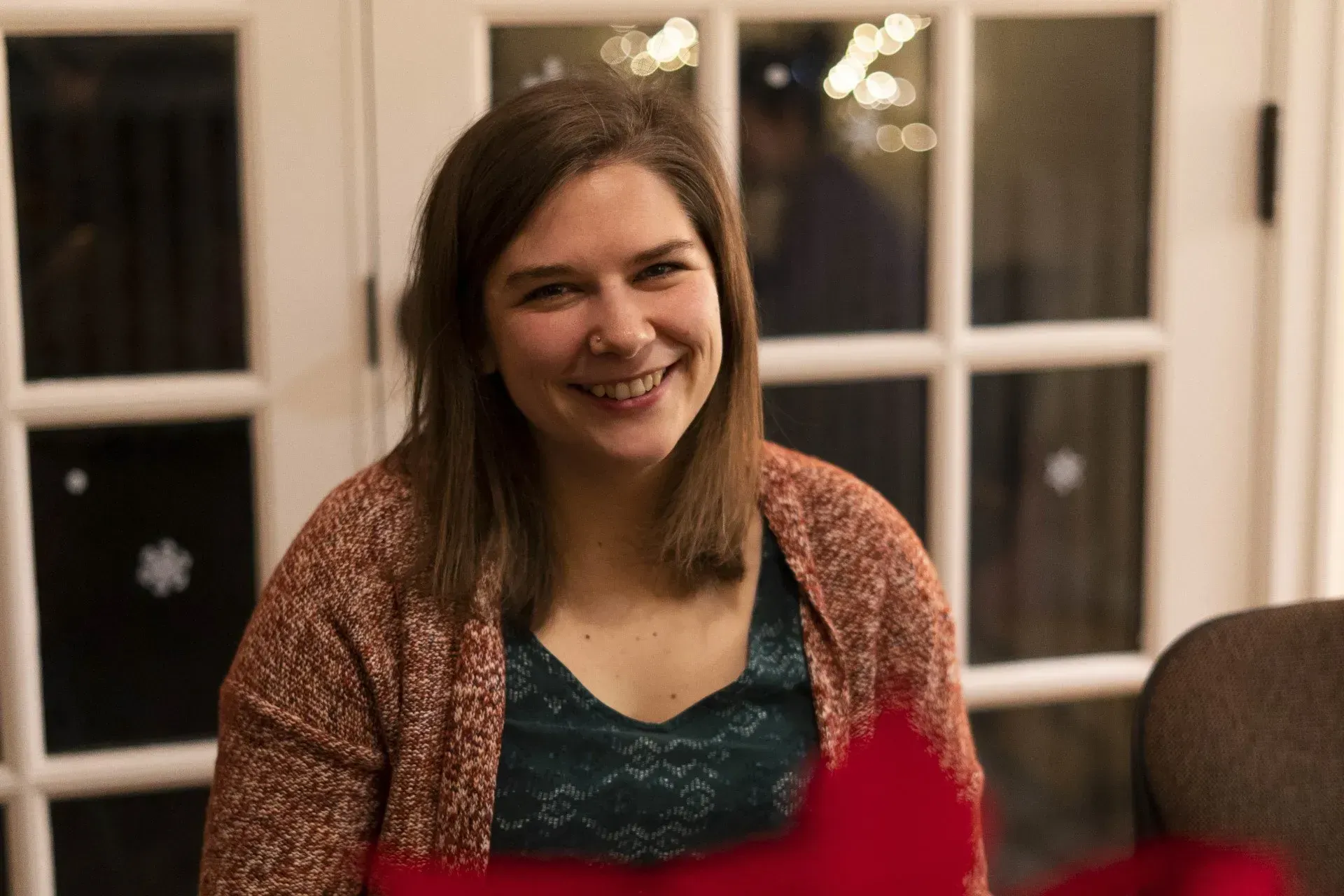 Woman with shoulder-length brown hair, wearing a red cardigan and green top, smiling in front of a window.