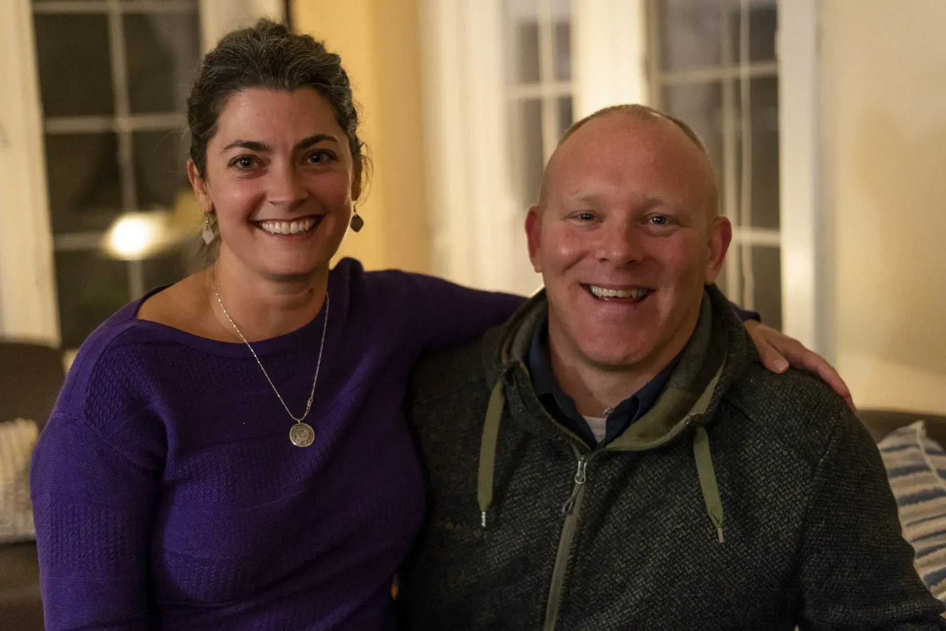 A smiling man and woman pose indoors. The woman wears a purple sweater, and the man has a green hoodie.
