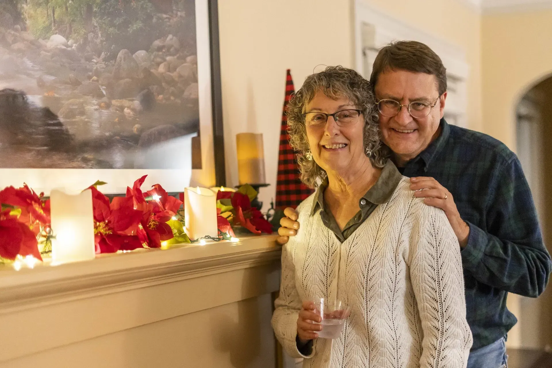 A smiling older couple poses by a decorated fireplace. 