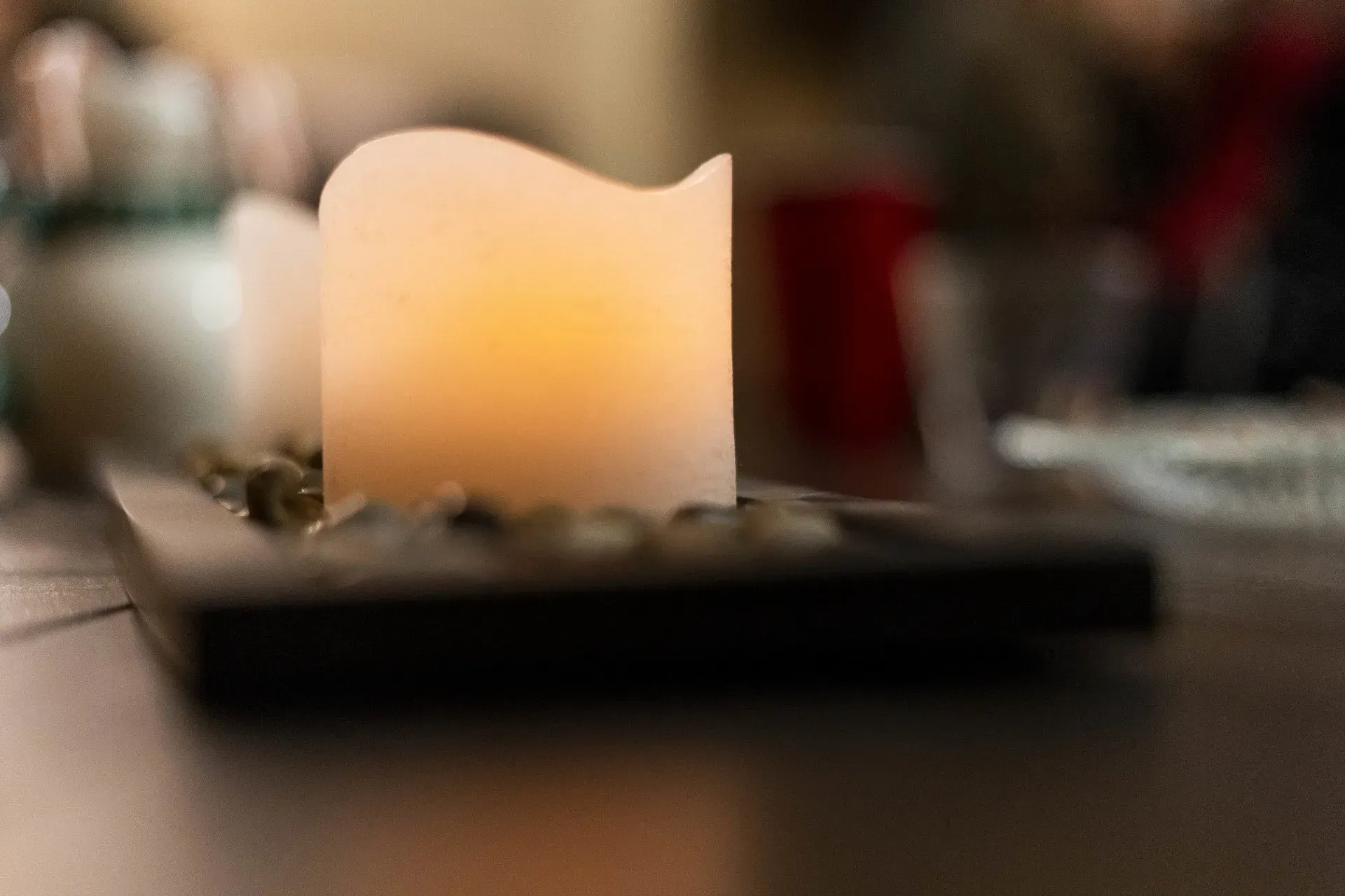 Glowing white candle in focus, set on a black rectangular base with small pebbles, in a dimly lit setting.