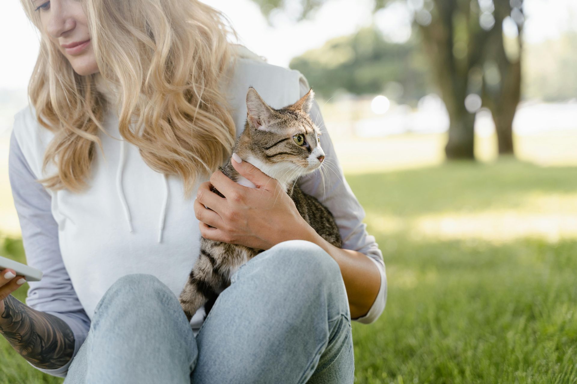 A woman is sitting on the grass holding a cat and looking at her phone.