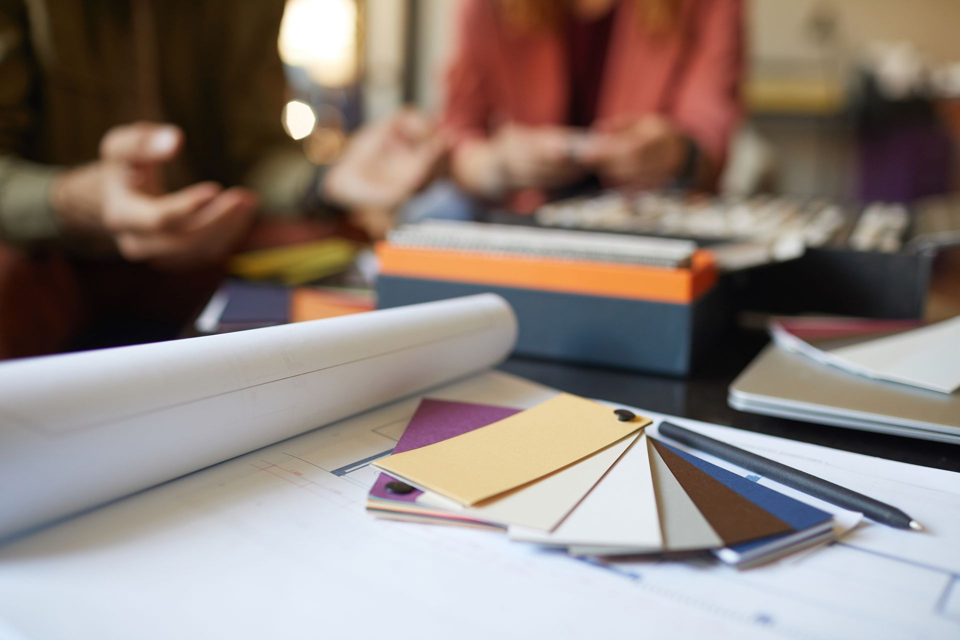 Designers collaborating over samples and blueprints; color swatches, pencil, and laptop on table.