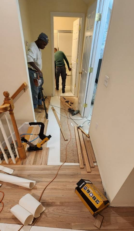 Construction in hallway with workers, tools, and materials. Wooden floors, tan walls.