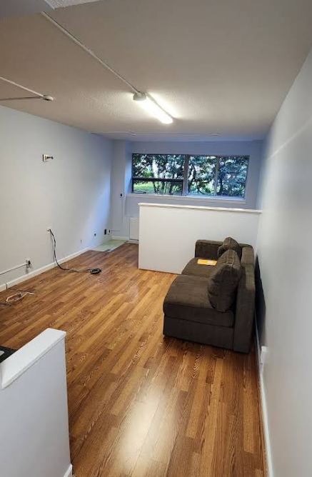A room with hardwood floors, white walls, and a brown sofa. A large window looks outside.