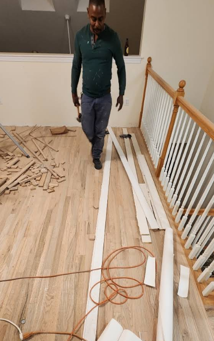 Man in green shirt, carrying hammer, on wood floor. Staircase and trim pieces visible.