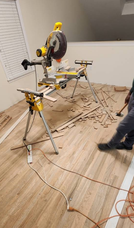 A miter saw on a stand, cutting wood for a hardwood floor installation. A person swings a hammer.