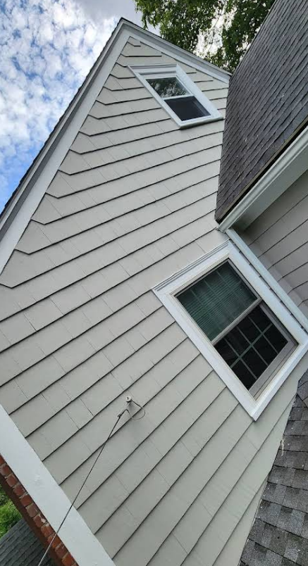 Exterior of a house with light gray siding, white trim, and two windows. A dark roof is visible on the right.