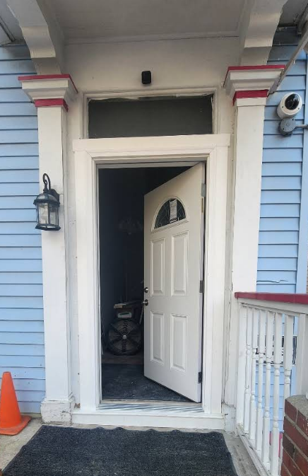 White door ajar, framed by white and red columns, exterior blue siding, security cameras.