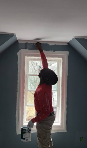 Person painting a room, reaching to paint a white ceiling trim above a window.