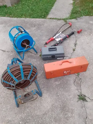 Two drain cleaning augers, two metal toolboxes, and two handheld drain tools arranged on a concrete driveway.