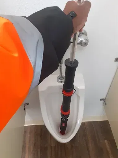 A person in a high-visibility orange jacket uses a manual plumbing tool to clear a clog in a white wall-mounted urinal.
