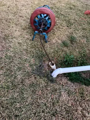 A drain cleaning machine with a red drum and blue stand feeds a metal snake cable into a white PVC pipe in the grass.