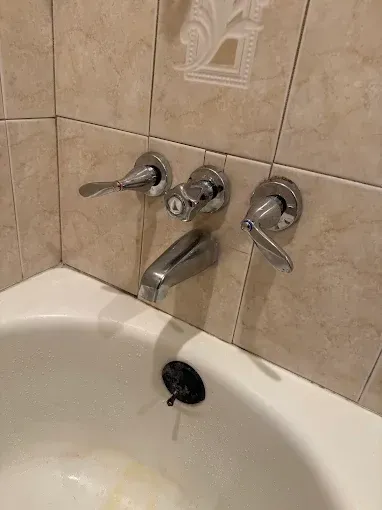 A chrome bathtub faucet assembly with two side handles, a central diverter knob, and a black overflow drain in a tiled wall.