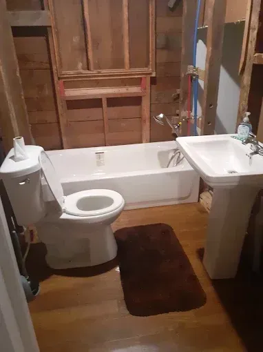A bathroom undergoing renovation featuring a white tub, toilet, and pedestal sink against exposed wooden wall studs.