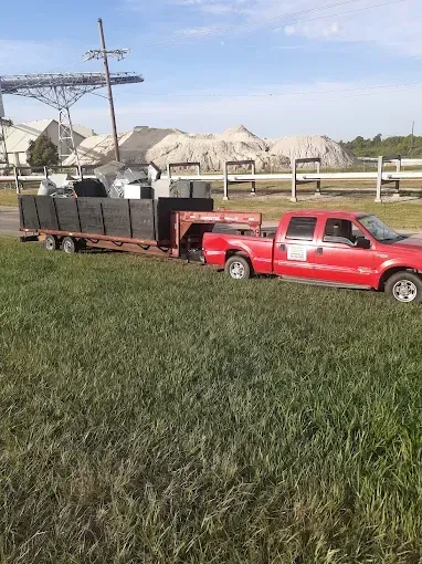 A red pickup truck pulling a trailer loaded with metal scraps and debris in a field near a large industrial facility.