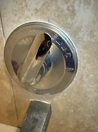 A close-up view of a metal shower faucet handle mounted on a tiled wall, showing a damaged, dark interior section.